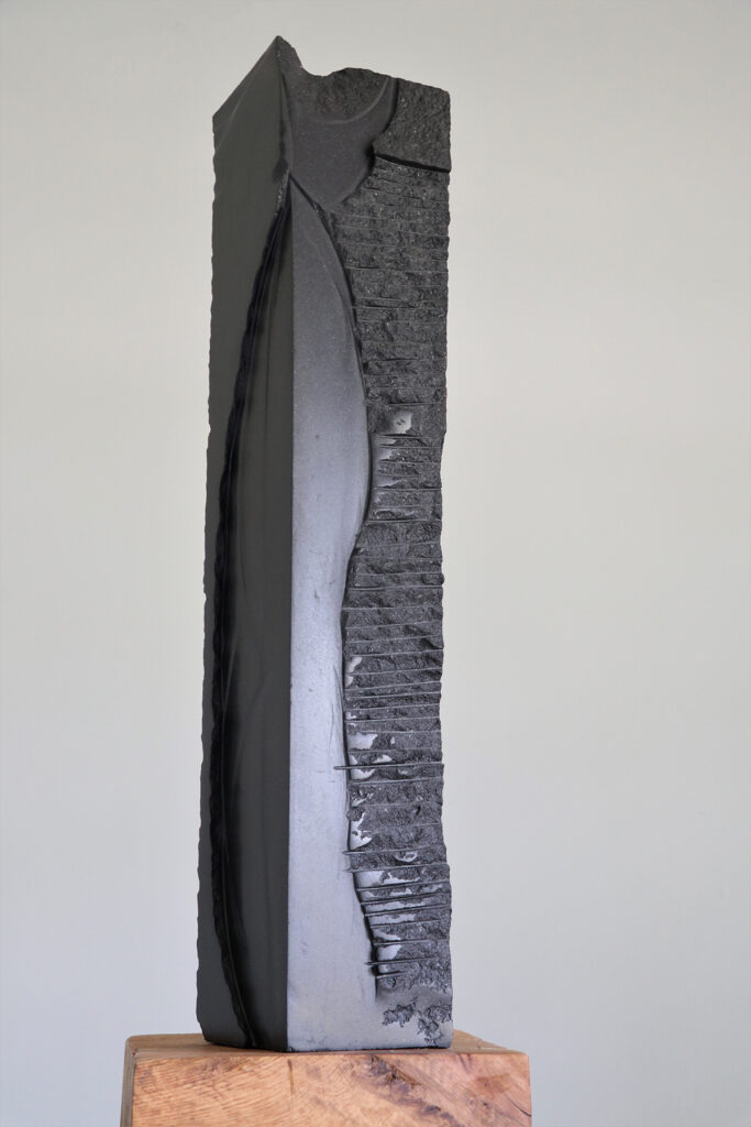 Tall, dark grey stone sculpture on wooden pedestal. 