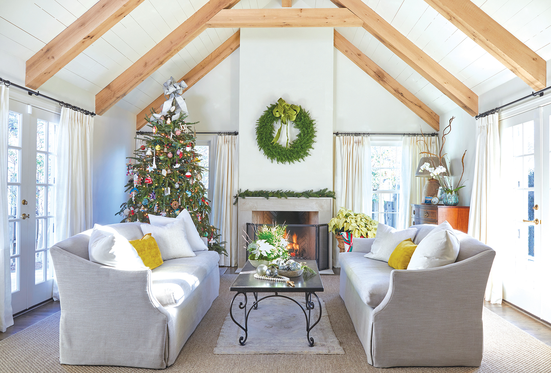 Living room with exposed beams decorated with a large wreath above the fireplace and Christmas tree. 
