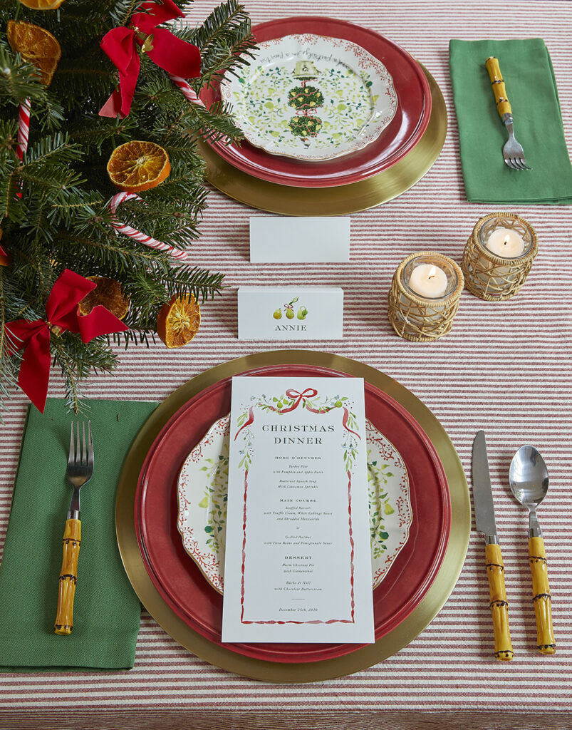 Detail photo of a table set with gold chargers, bamboo flatware, green napkins, and red Lenox dinner plates, along with a custom salad plate. Candleholders from Mary and Wilma and custom paper goods by Dogwood Hill featuring art by Sarah Robins Powell.