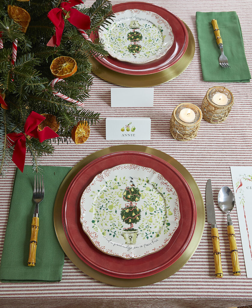 Alternate detail photo of a table set with gold chargers, bamboo flatware, green napkins, and red Lenox dinner plates, along with a custom salad plate. Candleholders from Mary and Wilma and custom paper goods by Dogwood Hill featuring art by Sarah Robins Powell.