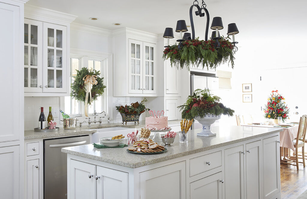 White kitchen with island covered in deserts. Greenery hanging from light fixture and wrath hung on the window above the sink.
