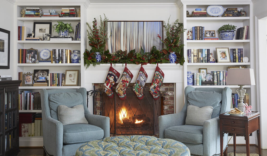 A lit fireplace is flanked by bookshelves. The mantel is styled with 4 stockings, red and green garland, and two antique blue and white vases.  