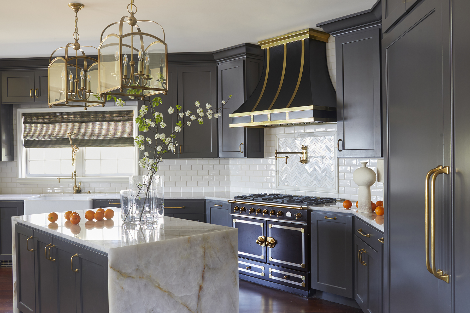Dark upper and lower kitchen cabinets with gold hardware, a black stove/over and matching range hood with gold accents contrasted by a white marble kitchen island with gold veins.